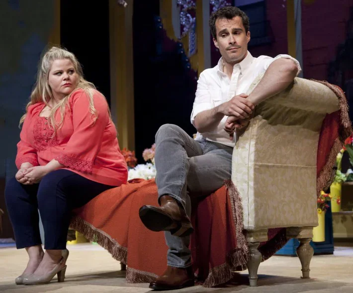 Deux acteurs se trouvent sur scène : une femme est assise sur une chaise et regarde un homme assis sur un canapé. Il est évident q