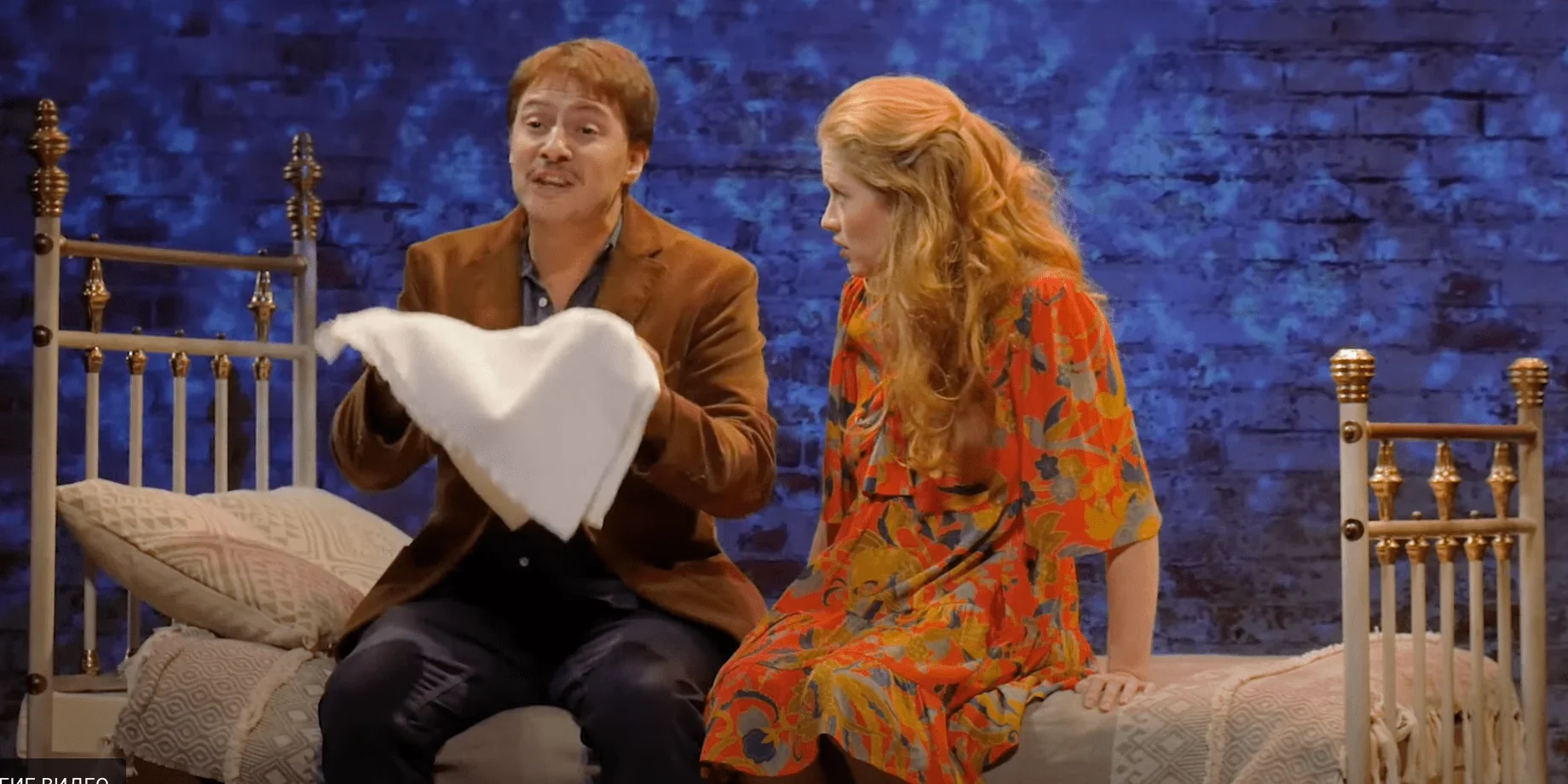 Sur scène, deux comédiens sont assis sur un lit. Un homme et une femme se regardent intensément, puis se tournent lentement vers l