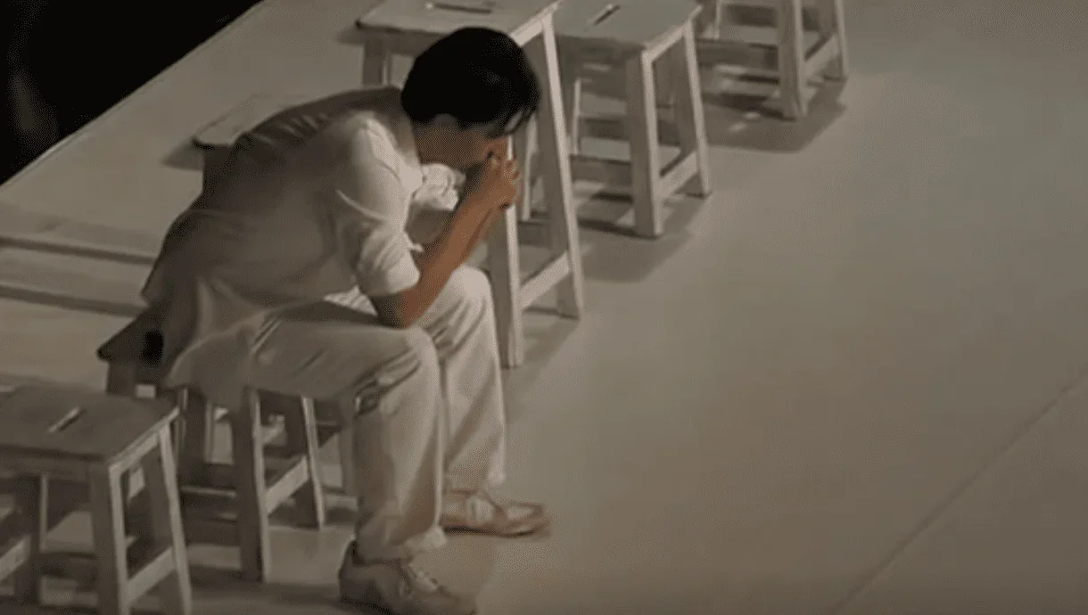 Un homme est assis sur une chaise, perdu dans ses pensées, son menton reposant dans ses mains, ses yeux fixés sur le sol.