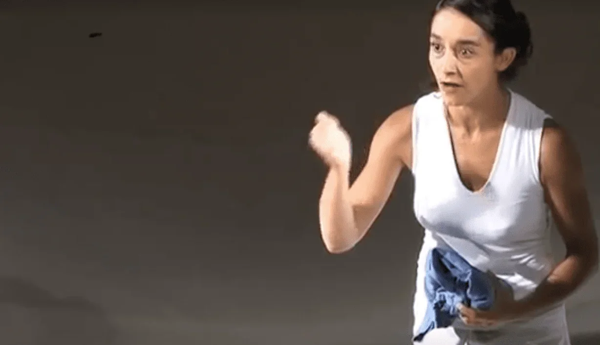 Sur scène, une femme vêtue d'une robe blanche captivante prononce son monologue avec une émotion palpable, s'adressant directement