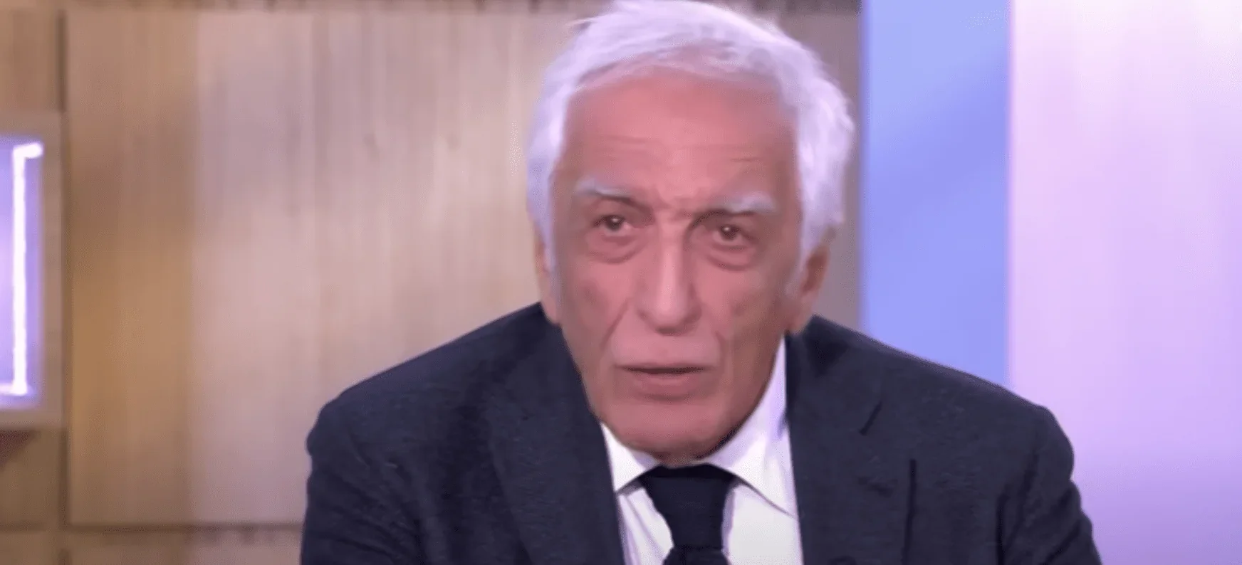 Gérard Darmon est assis confortablement dans un fauteuil, devant les caméras d'une émission de télévision. Il porte une chemise él