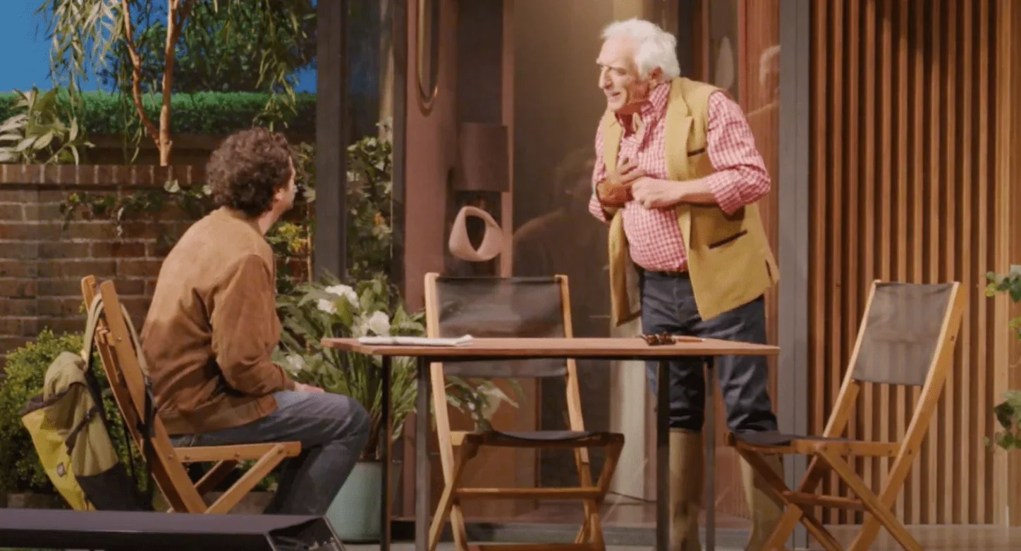 Gérard Darmon et Max Boublil se tiennent debout sur une scène, habillés en costumes élégants. Gérard Darmon semble calme et posé,