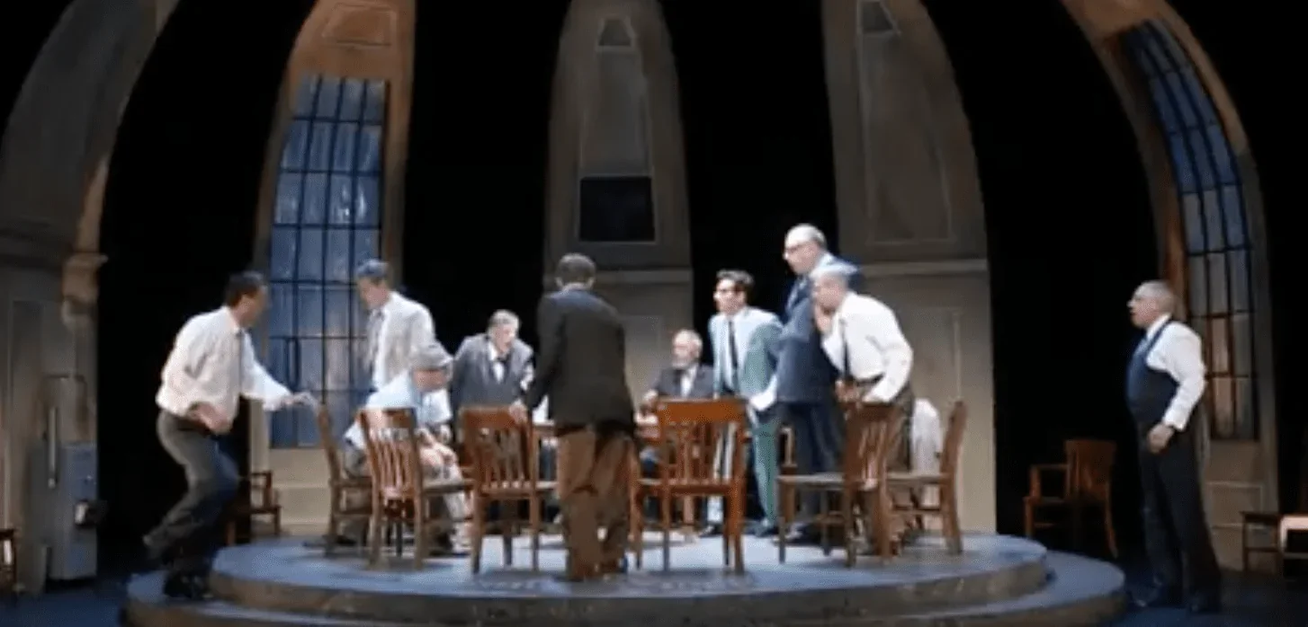 L'image capturée présente une scène emblématique du spectacle "12 hommes en colère". Sur scène, tous les personnages sont présents