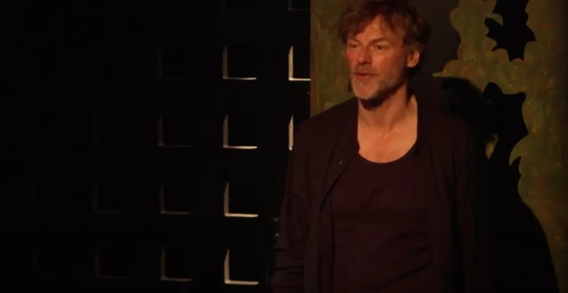 Marcello, sous un éclairage tamisé, se tient au centre de la scène, captivant l'audience par son récit. Son expression est intense