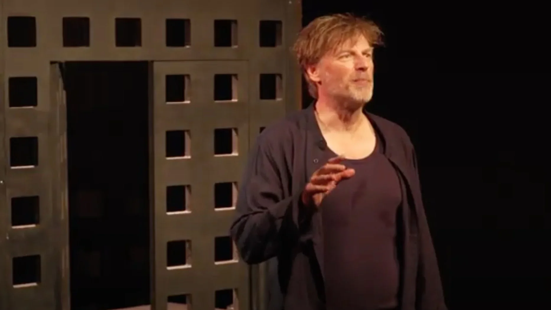 Marcello se tient debout sur scène, une figure solitaire dans la lumière. Il est droit, déterminé, un symbole de résistance et de