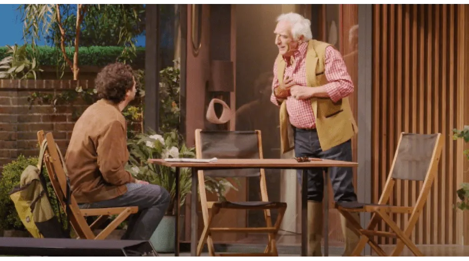 Gérard Darmon et Max Boublil se tiennent debout sur une scène, habillés en costumes élégants. Gérard Darmon semble calme et posé, 