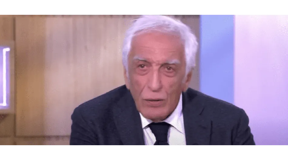 Gérard Darmon est assis confortablement dans un fauteuil, devant les caméras d'une émission de télévision. Il porte une chemise él